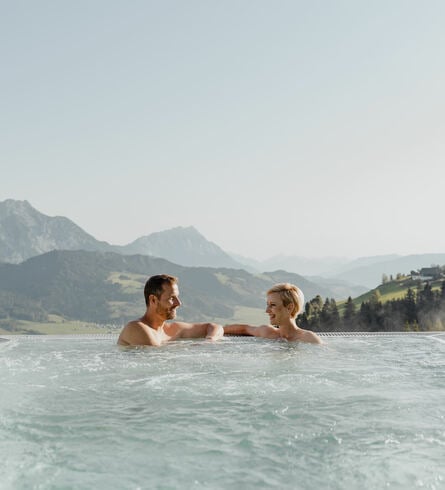 Paar unterhält sich im Luxury Infinity Pool des Natur- und Wellnesshotel Höflehner. Im Hintergrund erkennt man die Ennstaler Bergwelt.